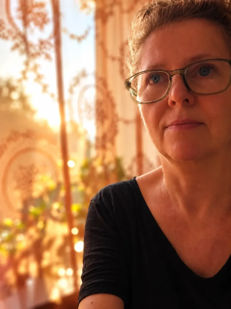 Monique bij het raam van haar praktijk, met warm zonlicht door het kanten gordijn